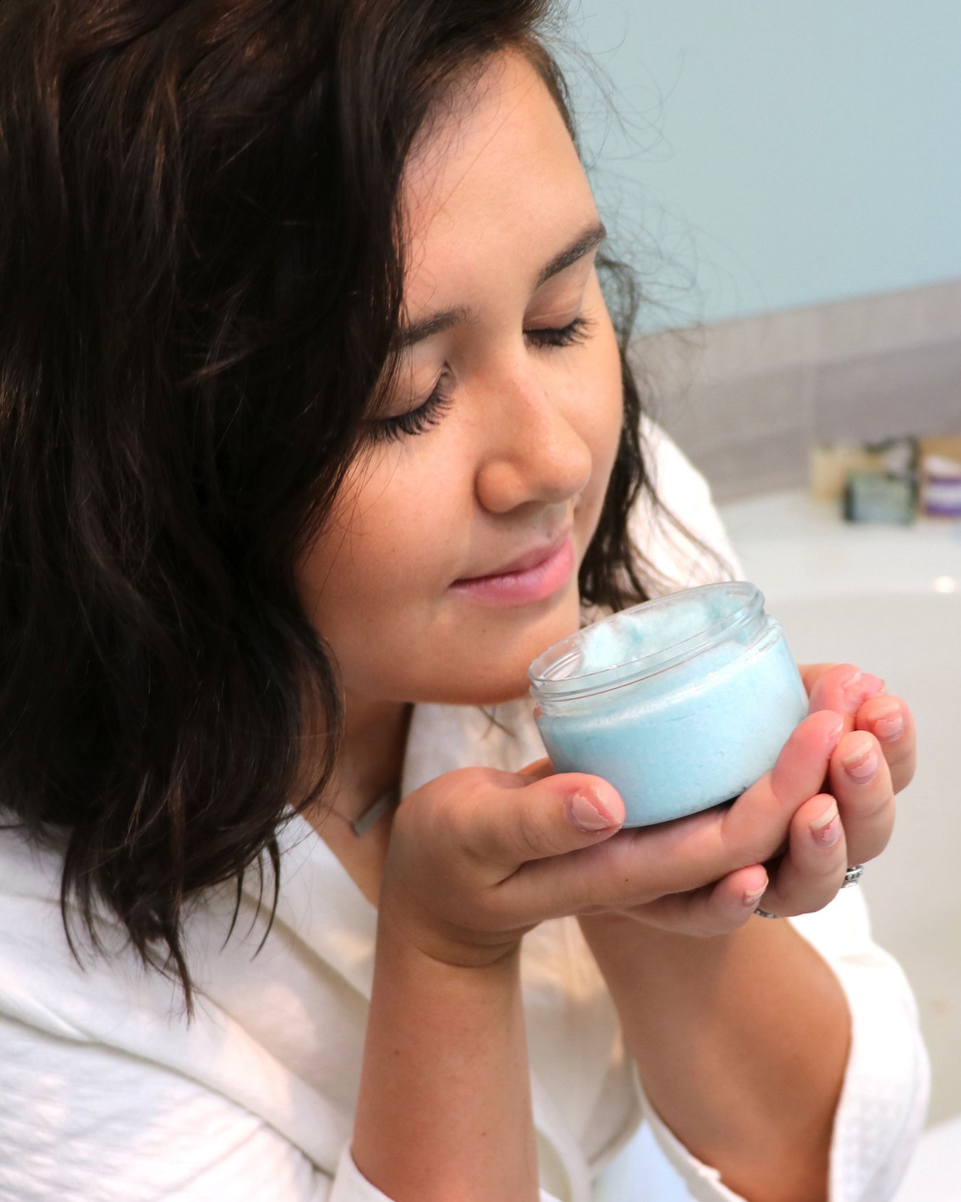 A woman with dark hair, wearing a white robe, holds a small clear jar filled with light blue cream near her face, smelling the product in a bathroom setting.