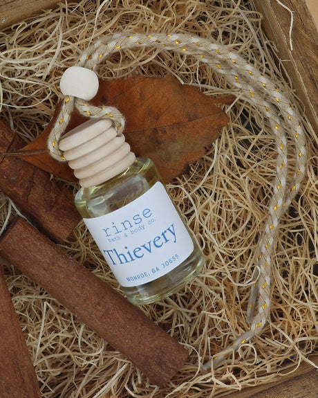 Thievery car diffuser with jute cord lying on excelsior beside cinnamon sticks and a dried leaf in a wooden crate
