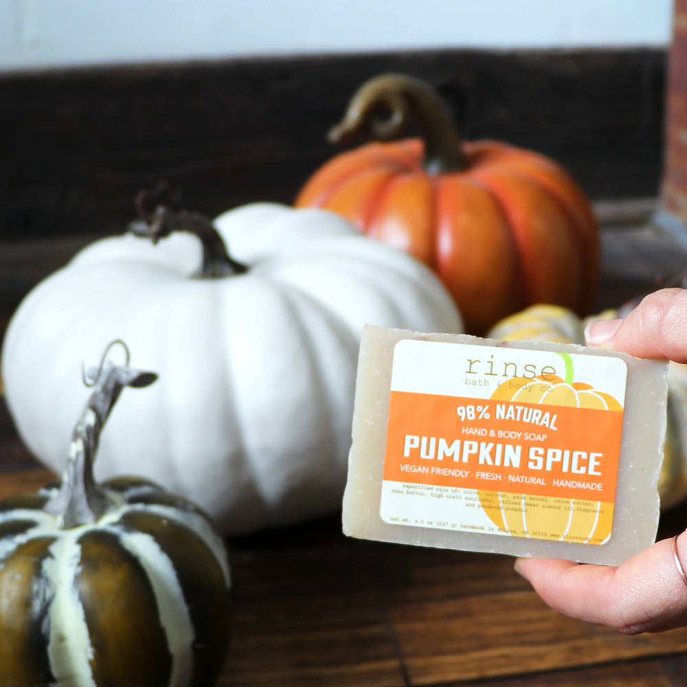 A hand holding the Pumpkin Spice bar in front of heirloom white and green pumpkins on weathered wood — nutmeg, clove and brown sugar, the bar that makes an ordinary shower smell like the first cold morning. Rinse Bath & Body Co.