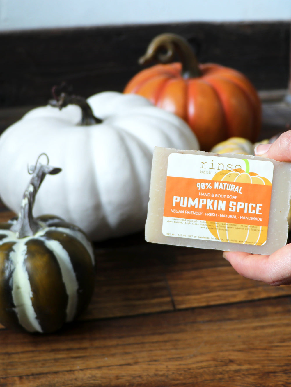 A hand holding the Pumpkin Spice bar in front of heirloom white and green pumpkins on weathered wood — nutmeg, clove and brown sugar, the bar that makes an ordinary shower smell like the first cold morning. Rinse Bath & Body Co.