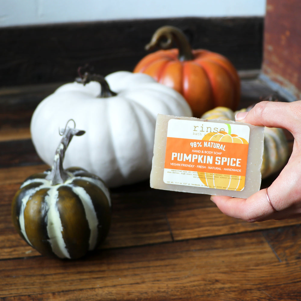 A hand holding the Pumpkin Spice bar in front of heirloom white and green pumpkins on weathered wood — the soap that anchors this fry-box gift set. Rinse Bath & Body Co.