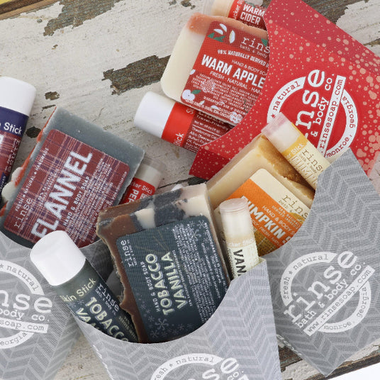A curated bath/body gift assortment from Rinse & Body Co. arranged in a gray paper tray on a distressed white wood surface.