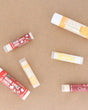 Warm Apple Cider lip balms scattered across warm kraft paper with the season's other flavours — spiced apple, clove and brown sugar for chapped autumn lips. Rinse Bath & Body Co.