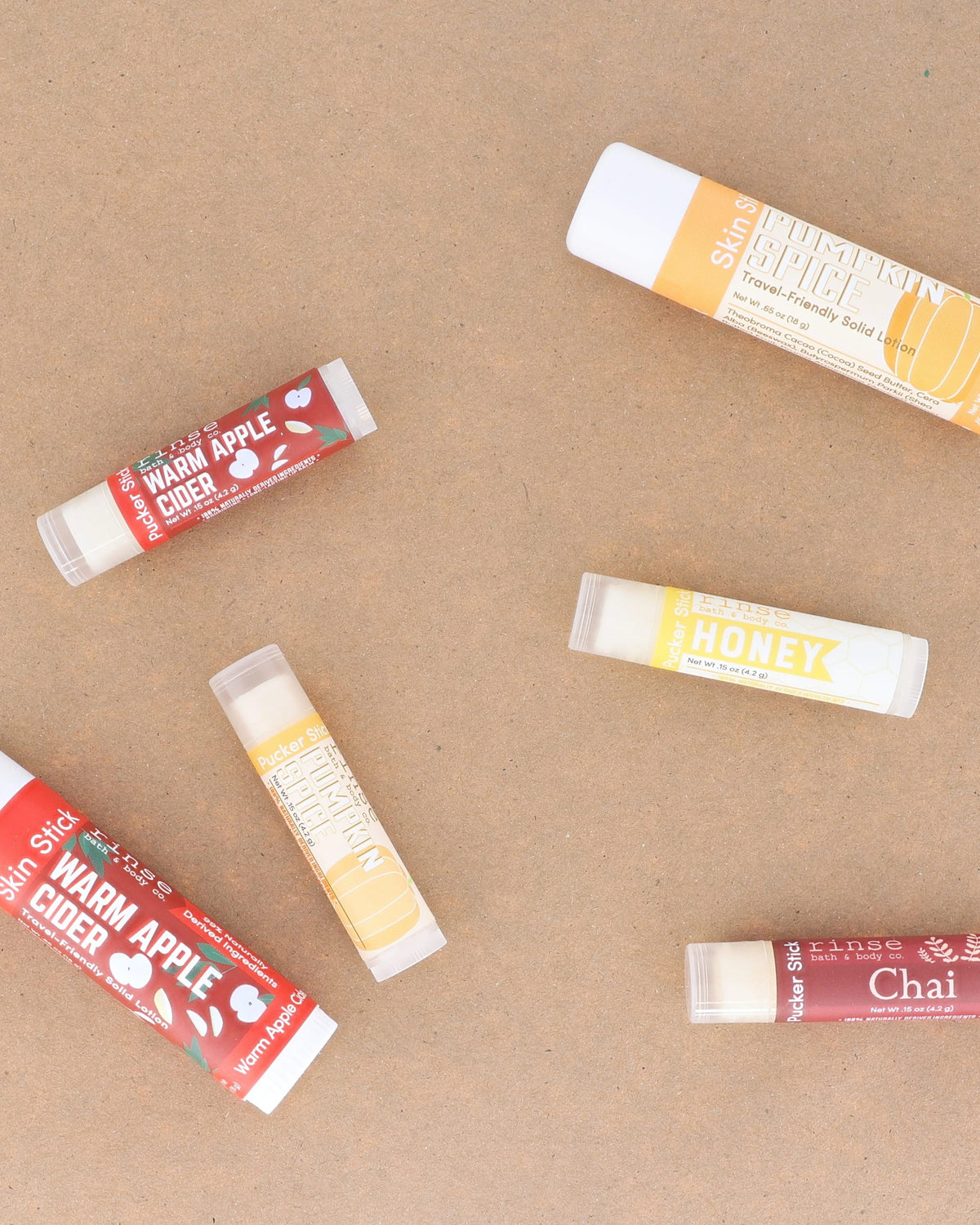 Warm Apple Cider lip balms scattered across warm kraft paper with the season's other flavours — spiced apple, clove and brown sugar for chapped autumn lips. Rinse Bath & Body Co.