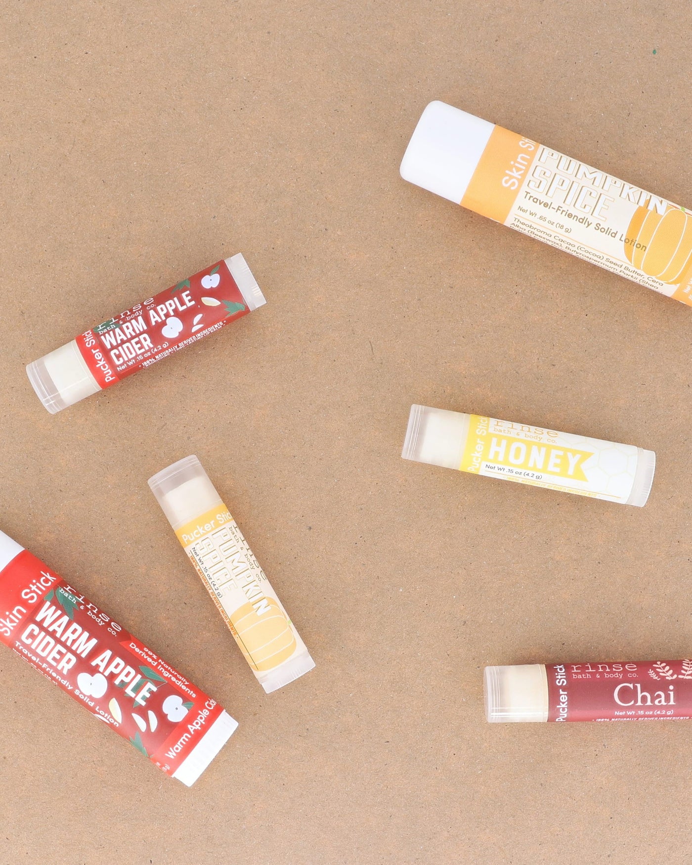 Warm Apple Cider lip balms scattered across warm kraft paper with the season's other flavours — spiced apple, clove and brown sugar for chapped autumn lips. Rinse Bath & Body Co.