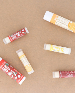 Warm Apple Cider lip balms scattered across warm kraft paper with the season's other flavours — spiced apple, clove and brown sugar for chapped autumn lips. Rinse Bath & Body Co.