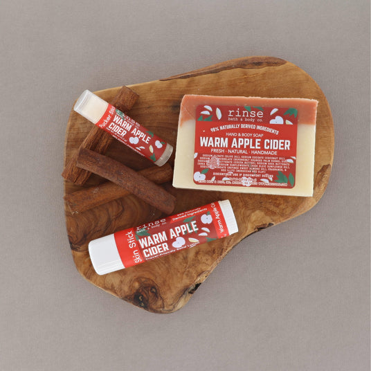 A set of warm apple cider scented products from the brand Rinse, including a bar of soap and two lip balm tubes, arranged on a rustic wooden slice over cinnamon sticks against a neutral gray background.