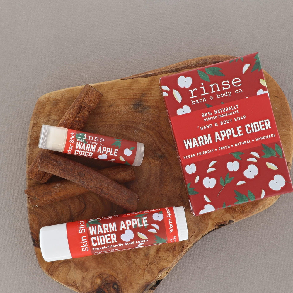 Two Warm Apple Cider solid lotion sticks resting on a live-edge walnut board beside their red harvest-print cartons — spiced apple, clove and brown sugar, made for the first cold morning of sweater weather. Rinse Bath & Body Co.