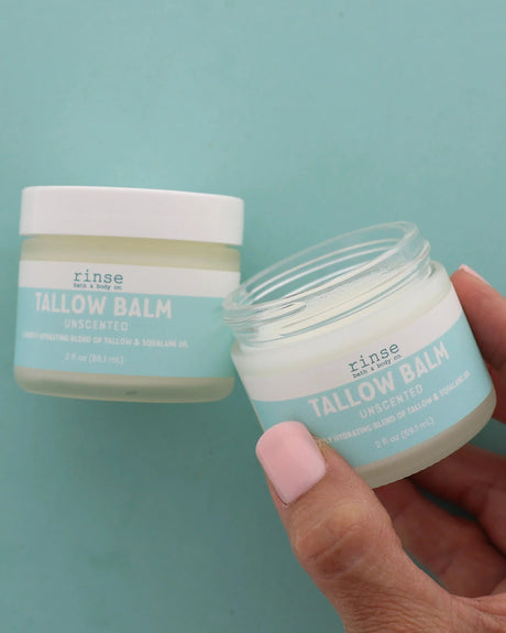 A hand holding an open jar of Unscented Tallow Balm with a second closed jar behind it, on a teal background. Rinse Bath & Body Co.