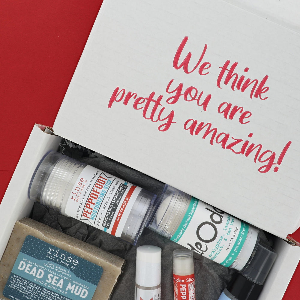 An open white gift box filled with bath and body items, photographed from above on a red background. The inside lid reads 'We think you are pretty amazing!' in pink script.