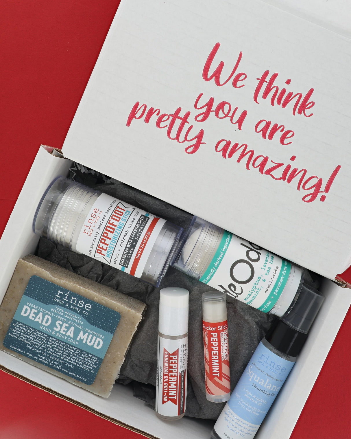 An open white gift box filled with bath and body items, photographed from above on a red background. The inside lid reads 'We think you are pretty amazing!' in pink script.