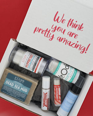 An open white gift box filled with bath and body items, photographed from above on a red background. The inside lid reads 'We think you are pretty amazing!' in pink script.