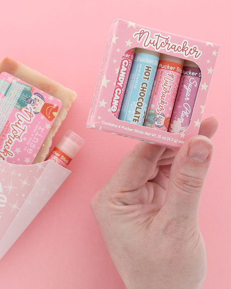 Hand holding the Nutcracker lip balm four-pack box on pink, with a pink Nutcracker gift set carton beside it