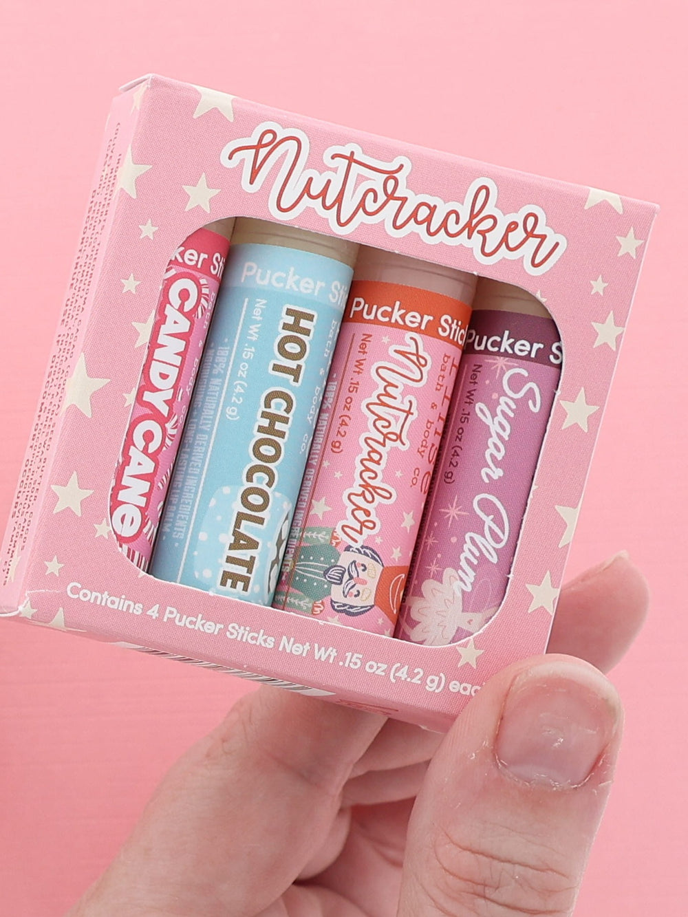 Hand holding the Nutcracker lip balm four-pack with Candy Cane, Hot Chocolate, Nutcracker and Sugar Plum sticks on pink
