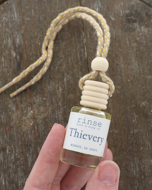 Hand holding a Thievery car diffuser by its glass bottle, wooden cap and jute cord over a dark wood board