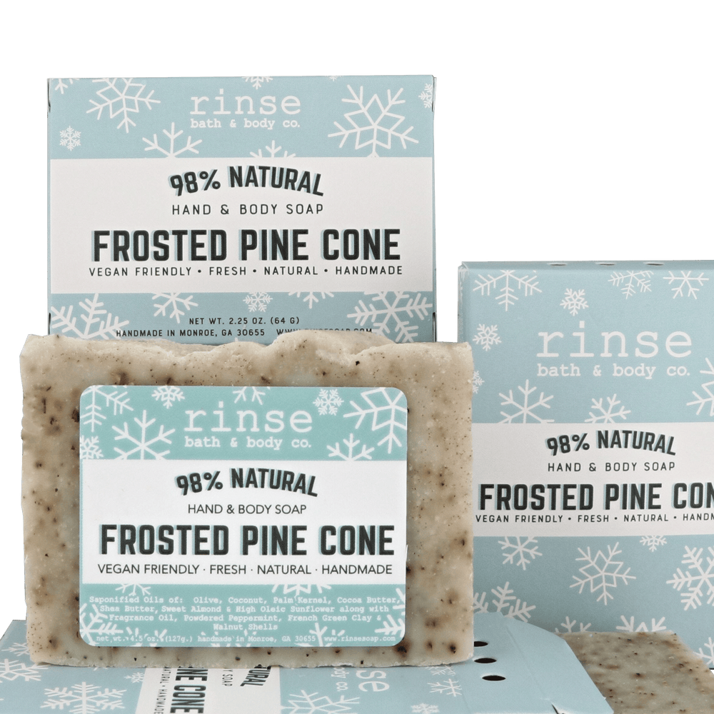 Mini Frosted Pine Cone soap standing before snowflake cartons on forest green — speckled cream bar in crisp winter light