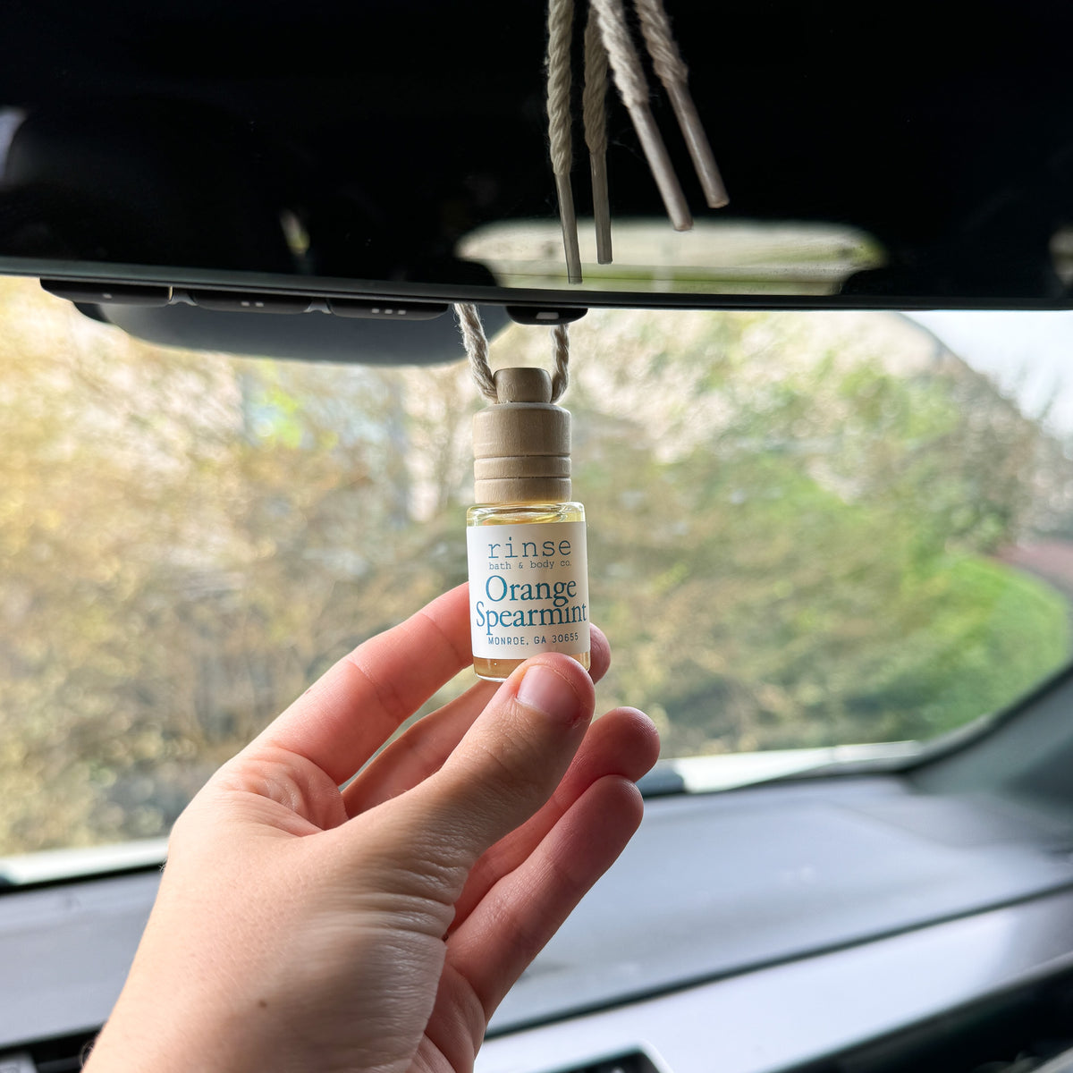 Orange Spearmint car diffuser hanging from the rearview mirror, steadied by hand in a sunlit car