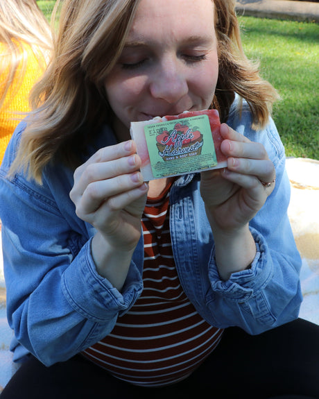 A woman on a picnic blanket lifting Apple Harvest soap to breathe it in, friends and autumn sunlight behind her — orchard-sweet, sweater-weather comfort. Rinse Bath & Body Co.