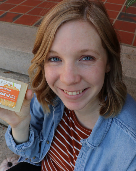 A woman outdoors smiling as she lifts a bar of Pumpkin Spice soap to breathe it in — warm autumn spice, the kind of scent you stop and notice. Rinse Bath & Body Co.