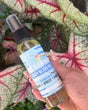 A hand holding a clear spray bottle of 'The Hawaiian Body Bliss Oil' in front of a background of green and red caladium leaves.