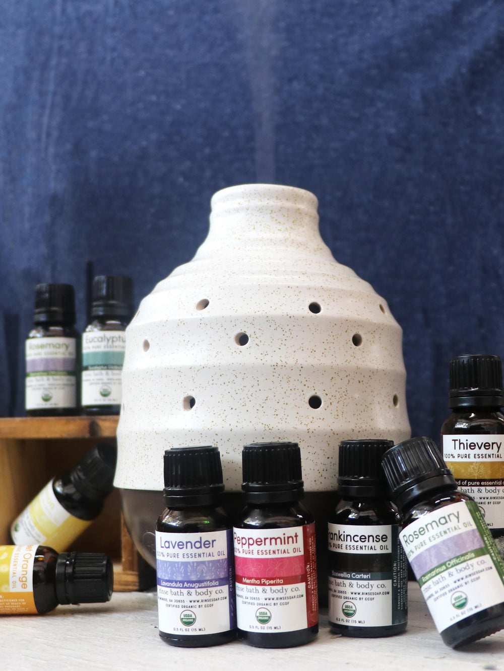 Lemon oil lying among Lavender Peppermint and friends around a ceramic diffuser — navy cloth and wood crate