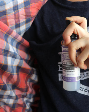 A hand holding a small bottle of ambient spray with a black cap and white label, positioned over a navy blue t-shirt and a red, blue, and white plaid blanket.