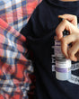 A hand holding a small bottle of ambient spray with a black cap and white label, positioned over a navy blue t-shirt and a red, blue, and white plaid blanket.