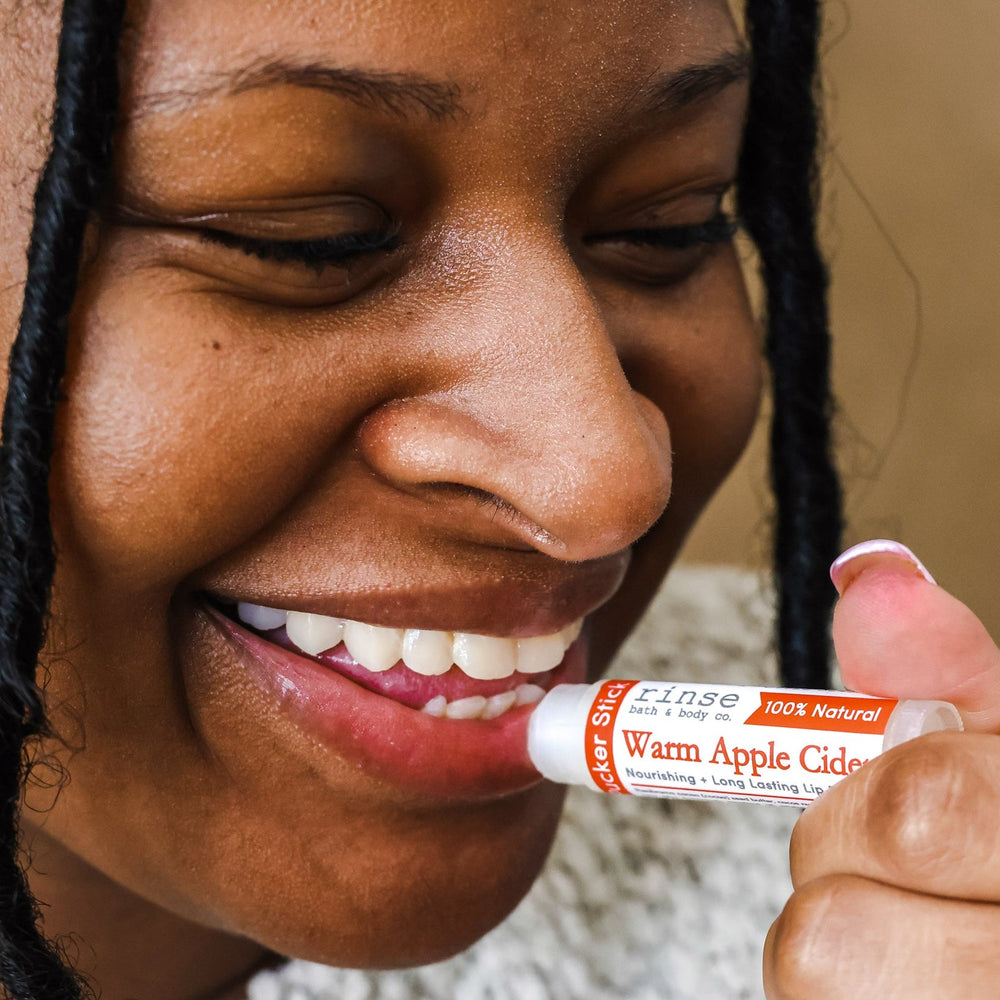 A woman smiling as she swipes on Warm Apple Cider lip balm, cable-knit sweater at her shoulder — cider-spiced comfort on the first cold day. Rinse Bath & Body Co.