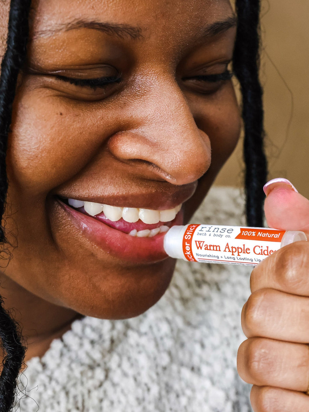A woman smiling as she swipes on Warm Apple Cider lip balm, cable-knit sweater at her shoulder — cider-spiced comfort on the first cold day. Rinse Bath & Body Co.