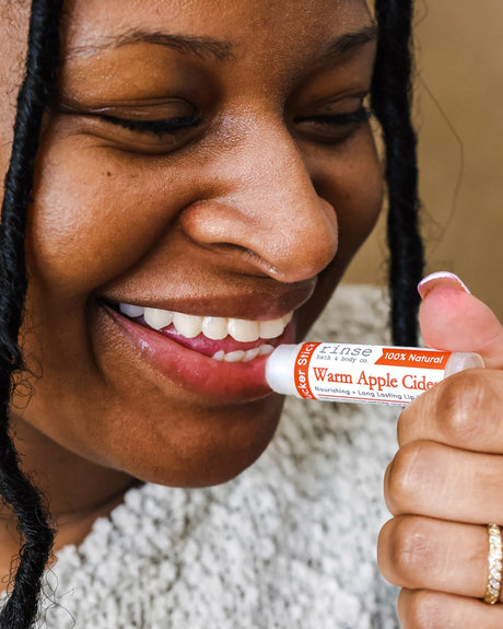 A woman smiling as she swipes on Warm Apple Cider lip balm, cable-knit sweater at her shoulder — cider-spiced comfort on the first cold day. Rinse Bath & Body Co.