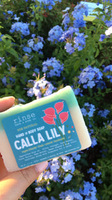 Hand presenting Calla Lily soap among plumbago blooms — sunlit garden greens behind