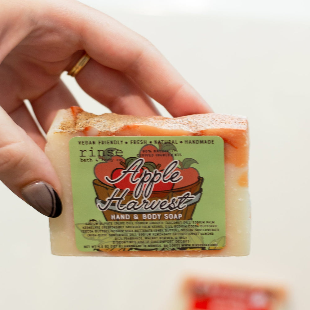A hand holding the Apple Harvest bar with its orchard-print label facing out — crisp red apple and warm cinnamon, the soap that makes an ordinary shower feel like a fall morning. Rinse Bath & Body Co.