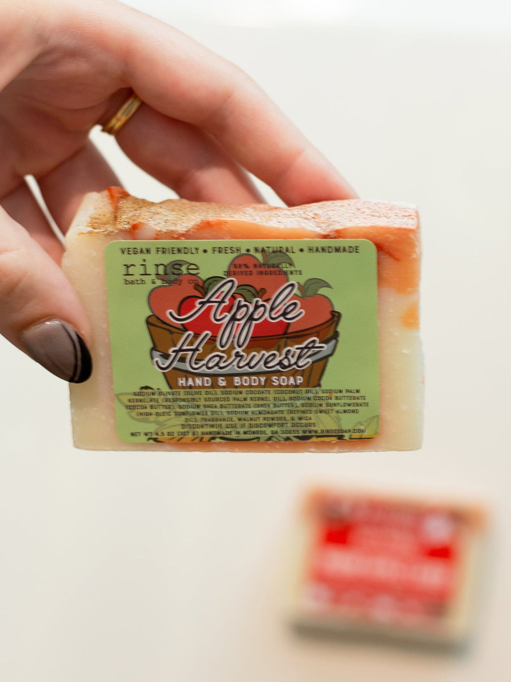 A hand holding the Apple Harvest bar with its orchard-print label facing out — crisp red apple and warm cinnamon, the soap that makes an ordinary shower feel like a fall morning. Rinse Bath & Body Co.