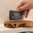 A hand setting the dark Tobacco Vanilla bar onto a live-edge wood board against warm tile, label facing out — smoky-sweet vanilla and cured tobacco leaf, a grown-up kind of cozy. Rinse Bath & Body Co.