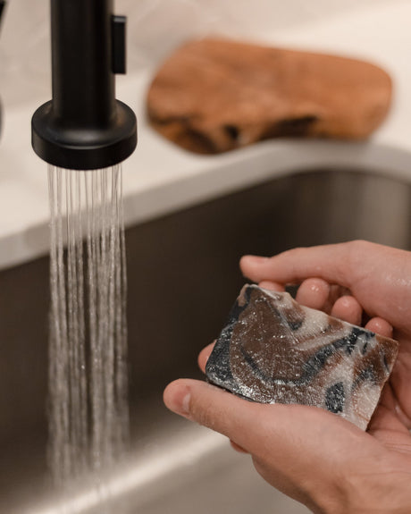 Tobacco Vanilla bar soap held in hand, its charcoal-dark top catching the light. Rinse Bath & Body Co.