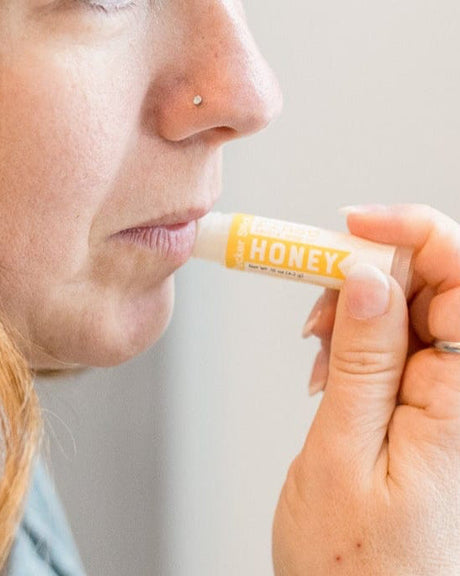 A woman swiping on Honey lip balm in soft daylight — raw honey and cocoa butter for lips that stay chapped. Rinse Bath & Body Co.
