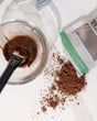 A mask brush resting in a bowl of mixed Cafe Mocha Face Mask, the pouch and loose coffee grounds beside it. Rinse Bath & Body Co.