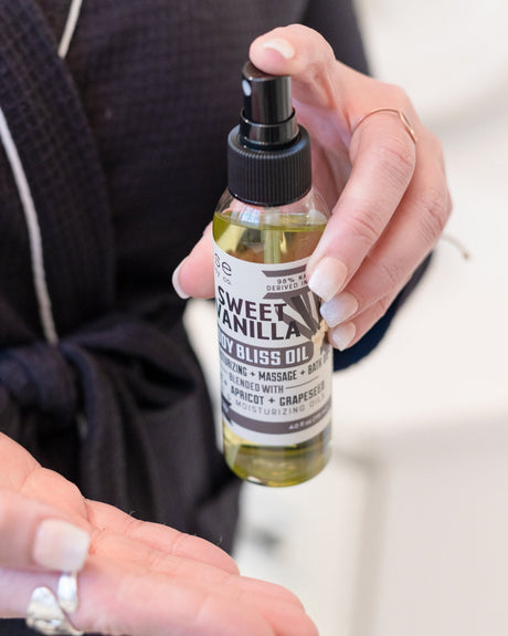 Sweet Vanilla Body Bliss oil in use, smoothed into skin — a fast-absorbing oil carrying warm bourbon vanilla. Rinse Bath & Body Co.