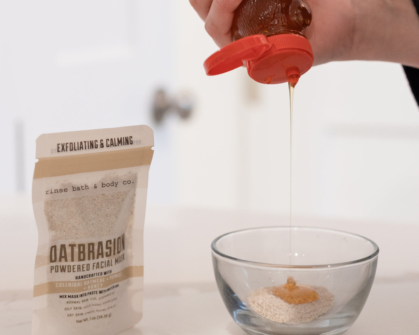 Honey poured from a bottle into a glass bowl of Oatbrasion mask powder, the pouch standing beside it — mix with water or honey into a paste. Rinse Bath & Body Co.