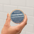 Two hands holding the round Dead Sea Mud Loofah bar with its blue label facing the camera — mineral mud and a loofah face for exfoliating and detoxing in one bar. Rinse Bath & Body Co.