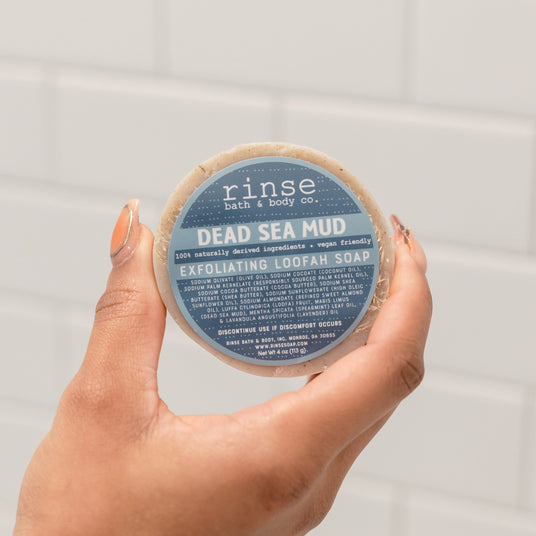 Two hands holding the round Dead Sea Mud Loofah bar with its blue label facing the camera — mineral mud and a loofah face for exfoliating and detoxing in one bar. Rinse Bath & Body Co.