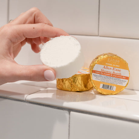 A hand holding a white Aller-Ease shower steamer puck beside its gold foil wrapper on white subway tile — one of the four in the box, lavender, lemon and peppermint steam for hay-fever season. Rinse Bath & Body Co.