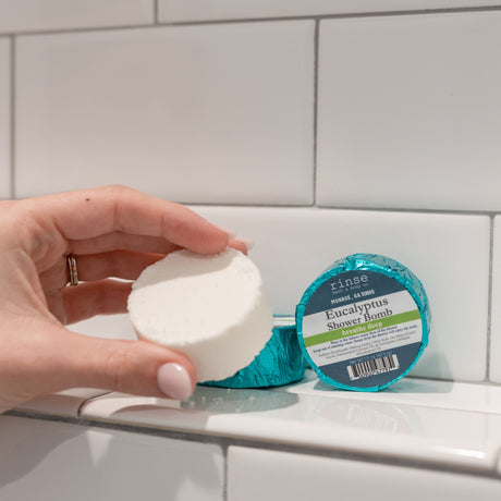 A hand holding a white Eucalyptus shower steamer puck beside its teal foil wrapper on a white-tiled shower ledge — one of the four in the box, steam-activated eucalyptus for a blocked-up morning. Rinse Bath & Body Co.