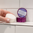 A hand holding a white Lavender shower steamer puck beside its purple foil wrapper on white subway tile — Bulgarian lavender released by shower steam. Rinse Bath & Body Co.