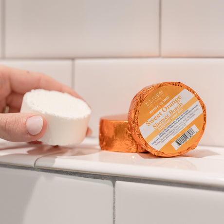 A hand holding a white Sweet Orange shower steamer puck beside its orange foil wrapper on white subway tile — one of the four in the box, bright citrus steam for a slow morning. Rinse Bath & Body Co.