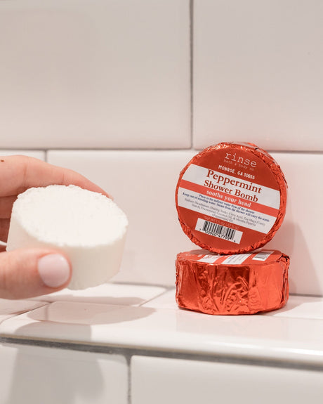 A hand holding a red foil-wrapped Peppermint shower steamer against white subway tile — set it on a ledge out of the stream and the steam carries the peppermint. Rinse Bath & Body Co.
