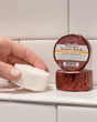 A hand holding a white Thievery shower steamer puck beside its maroon foil wrapper on white subway tile