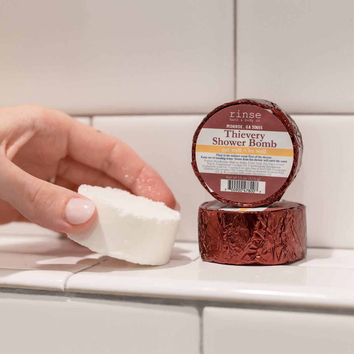 A hand holding a white Thievery shower steamer puck beside its maroon foil wrapper on white subway tile — one of the four in the box, warm cinnamon, clove and eucalyptus steam for cold season. Rinse Bath & Body Co.