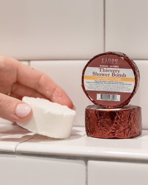 A hand holding a white Thievery shower steamer puck beside its maroon foil wrapper on white subway tile — warm cinnamon, clove and eucalyptus steam for cold season. Rinse Bath & Body Co.