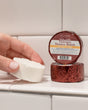A hand holding a white Thievery shower steamer puck beside its maroon foil wrapper on white subway tile — warm cinnamon, clove and eucalyptus steam for cold season. Rinse Bath & Body Co.
